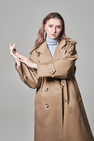 Attractive confident girl fashion model posing in a fashionable beige trench coat on grey studio background. Women's fashion. Autumn-spring.の写真素材