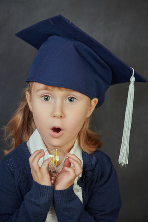 Education and early childhood development. Happy little student girl wearing academic hat looks to the camera very surprised. Background with black school board.の写真素材