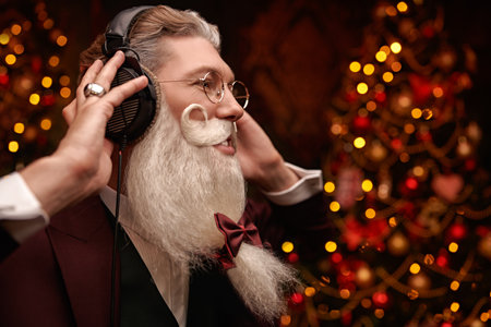 Portrait of a modern Santa Claus in headphones in an elegant suit against festive golden lights. Men's style in hair styling and beard, barbershop. Merry Christmas and New Year celebration.の写真素材