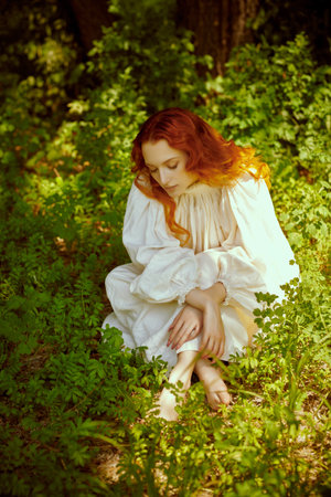 A beautiful romantic girl with charming wavy red hair, dressed in a long white shirt, rests in a park on a sunny summer day. The era of romanticism, 18-19 centuries.の写真素材