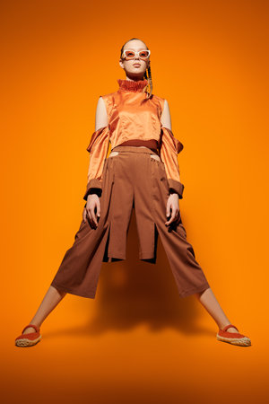 Fashion model girl posing in an orange top and brown wide-legs pants on an orange studio background. Summer haute couture collection.の写真素材