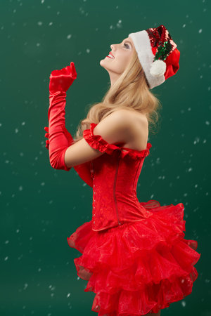 An attractive Santa girl with beautiful blond hair, dressed in a red outfit, admires the snowfall and smiles happily. Green studio background. Christmas and New Year concept.の写真素材