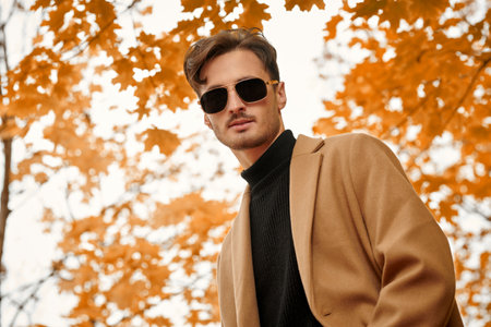 People and lifestyle. Autumn fashion. A handsome young man in a brown coat stands in an autumn park with a maple leaf in his hand and looks to the camera.の写真素材
