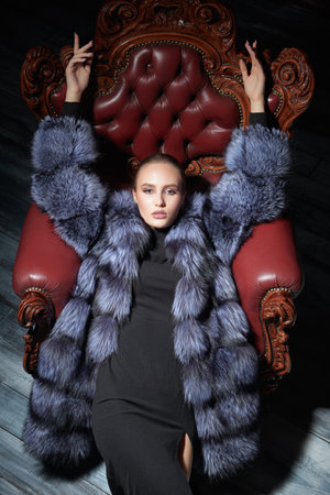 A chic blonde woman in an expensive silver fox fur coat sits in a vintage chair. Black background. Luxurious lifestyle of lady. Fur coat fashion.の写真素材