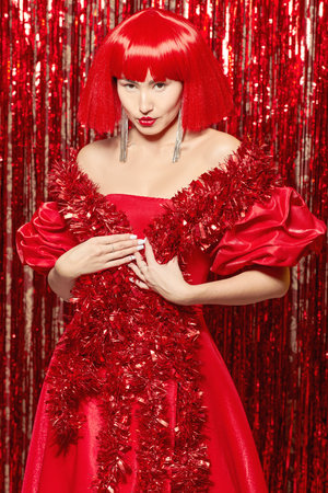 Stunning Asian lady model posing in an elegant red evening dress with puffs and a red wig against the backdrop of shiny foil curtains. Holiday fashion. Christmas and New Year.の写真素材