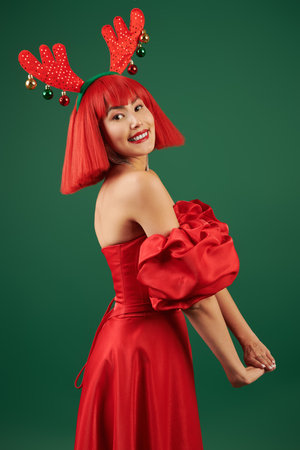 Celebration of Christmas and New Year. A pretty Asian girl model in an elegant red dress, a red wig and deer horns poses joyfully against a green background.の写真素材