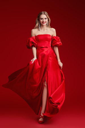 Evening Fashion. Full-length portrait of a gorgeous lady with lush wavy blonde hair posing in an elegant red evening dress with a neckline. Red studio background. Evening makeup and hairstyle. Luxury.の写真素材