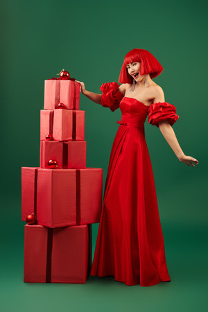 Festive and evening fashion. Beautiful Asian girl model in a chic red long dress, high heel shoes and a red wig poses with gift boxes on a green studio background. Full-length portrait.の写真素材
