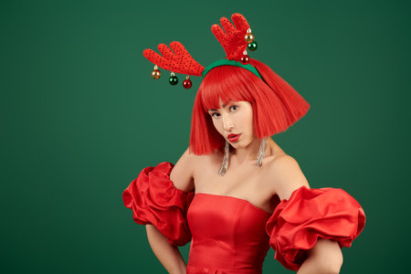 A wonderful celebration of Christmas and New Year. An attractive Asian girl model in an evening red dress, a red wig and deer horns poses against a green background. Copy space.の写真素材