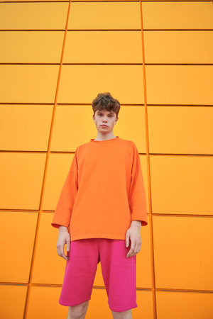 Portrait of a handsome young man in an orange sweatshirt and pink shorts against the background of a yellow building wall. Youth bright fashion style.の写真素材