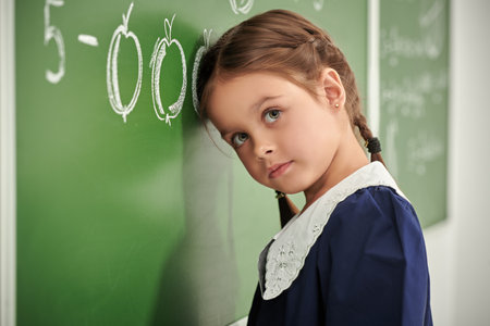 A little schoolgirl girl solves difficult tasks at the blackboard and looks thoughtful. Early development and education.の写真素材