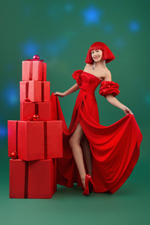Presents time. Beautiful Asian girl poses in a chic red long dress, high heel shoes and a red wig stands by gift boxes on a green studio background. Full-length portrait. Festive and evening fashion.の写真素材
