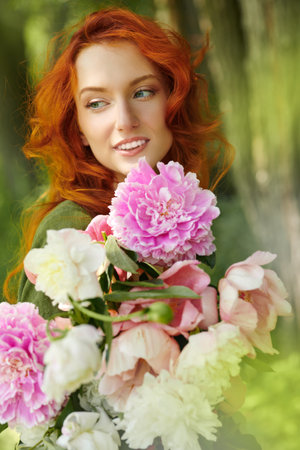 A charming red-haired girl stands with a large bouquet of peonies against the background of summer greenery. Women's beauty and flowers.の写真素材