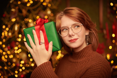 Merry Christmas and Happy New Year! A happy red-haired girl stands with a gift in her hands in a beautiful Christmas setting and smiles joyfully.の写真素材