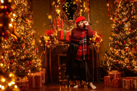 Festive Christmas and New Year. A cheerful pretty red-haired girl in a red beret stands with a gift in a fabulous Christmas room.の写真素材