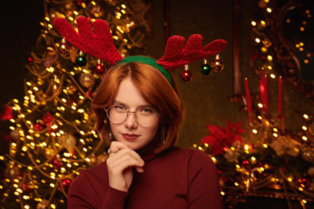 Portrait of a cute red-haired girl in deer antlers standing in a beautiful Christmas setting. New Year and Christmas celebration.の写真素材