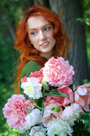 Portrait of a romantic red-haired lady standing with a large bouquet of peonies against the background of summer greenery. Women's beauty and flowers.の写真素材