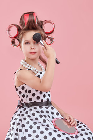 A little pretty girl in an elegant polka-dot dress and curlers on her head is going to a party and doing her make-up. Pink studio background. Kid's Fashion - clothes and accessories. Pin-up style.の写真素材