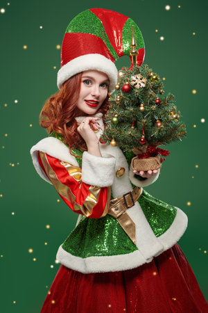 Merry beautiful Christmas fairy elf girl in a bright costume holds a christmas tree and smiles on a green studio background. Tales for Christmas and New Year.の写真素材
