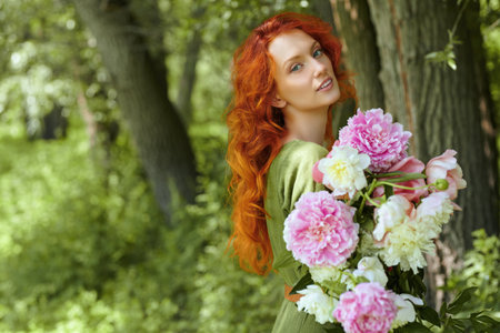 Female beauty and flowers. Portrait of a romantic red-haired young woman walking with a large bouquet of peonies in a summer park. Copy space.の写真素材