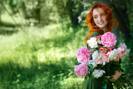 A charming red-haired girl stands with a large bouquet of peonies against the background of summer greenery. Women's beauty and flowers. Fantasy heroine. Celtic culture. Place for text.の写真素材