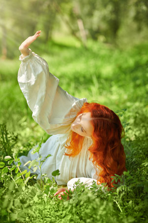 A beautiful girl with bright red wavy hair, dressed in a white nightgown, lies on the grass on a meadow and looks at the sun. The heroine of the novel. The era of romanticism, 18-19 centuries.の写真素材