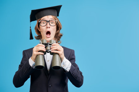 Education. A teenage student boy looks through binoculars and is very surprised. Modern generation. Studio blue background with copy space.の写真素材