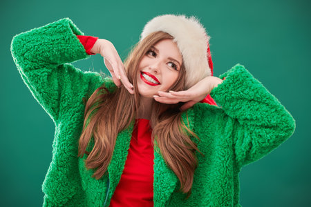 Merry Christmas and New Year! Happy cute girl with a charming smile posing in a fashionable green fur coat and Santa Claus hat. Green studio background.の写真素材