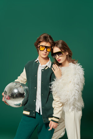 Fashion for Christmas and New Year. Stylish young guy and girl in fashionable outfits and sunglasses posing with disco balls on a green studio background. Copy space. Party style.の写真素材