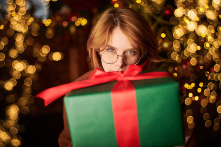 Merry Christmas! Happy New Year! A happy red-haired girl stands with a gift in her hands in a beautiful Christmas setting and smiles joyfully.の写真素材