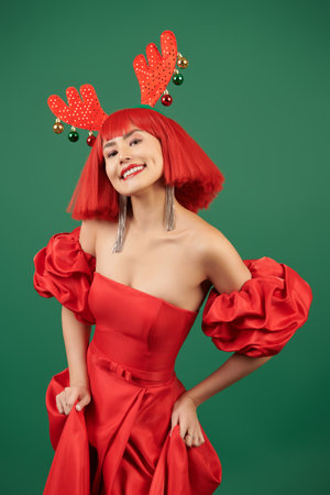 Celebration of Christmas and New Year. A pretty Asian girl model in an elegant red dress, a red wig and deer horns poses joyfully against a green background.の写真素材