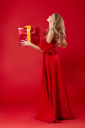 Christmas Fashion and Beauty. Full-length portrait of a stunning young woman with beautiful blonde hair, posing in a long elegant red dress and a gift. Red studio background.の写真素材