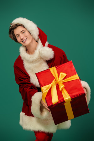A gift from Santa Claus for Christmas and New Year! Handsome teenager boy dressed in Santa Claus costume holds a gift box on a green studio background.の写真素材