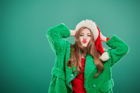 Portrait of a cute hipster girl in a bright green fur coat and Santa Claus hat posing on a green studio background. Copy space. Christmas and New Year concept.の写真素材