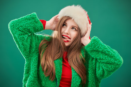 A cute hipster girl in a bright green fur coat and Santa Claus hat posing on a green studio background. Christmas and New Year concept.の写真素材