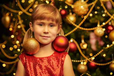 Children's Christmas and New Year. A cute little girl in a festive dress stands next to a Christmas tree and smiles.の写真素材