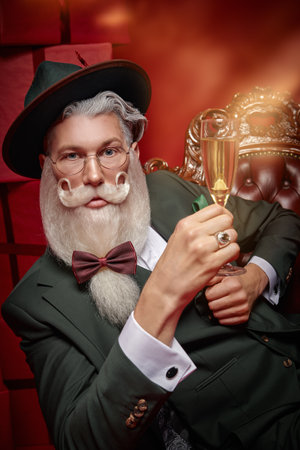 An elegant Santa Claus with a beautiful festive beard and mustache  in a stylish green suit holds a glass of champagne. Red background. Christmas, New Year.の写真素材