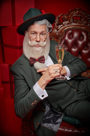An elegant Santa Claus with a beautiful festive beard and mustache  in a stylish green suit holds a glass of champagne. Red background. Christmas, New Year.の写真素材
