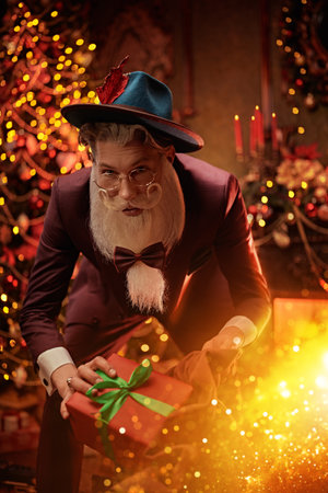 Gift from stylish Santa Claus. Handsome modern Santa Claus in an elegant festive costume gives a gift and smiles joyfully while standing in a magical festive setting at home. Christmas and New Year.の写真素材