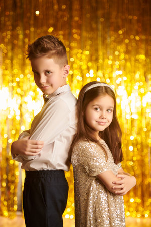 Children's holiday fashion. Two beautiful children - a boy and a girl - pose together in evening dresses against shiny gold foil curtains. Christmas and New Year.の写真素材