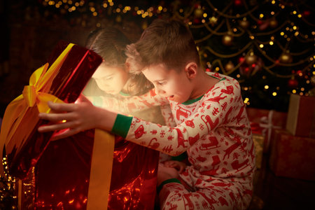 Magical Christmas and New Year. Two children in pajamas open a gift while sitting near the Christmas tree and are surprised.の写真素材