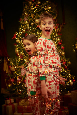 Two funny children in pajamas are standing at home near the Christmas tree, tied with a garland and are screaming in shock. Crazy Christmas and New Year.の写真素材
