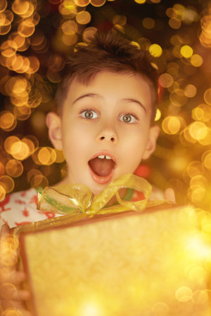Happy boy holding a gift in his hands and rejoicing, surrounded by golden holiday lights. Christmas and New Year.の写真素材
