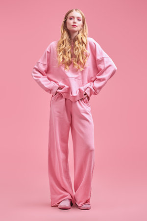 Beautiful girl fashion model with long wavy blond hair posing in a pink sweatshirt and trousers on a pink studio background. Modern youth feminine style.の写真素材