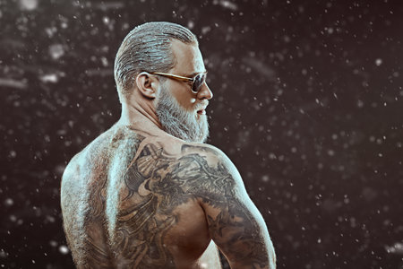 A handsome brutal muscular man with a white beard and hair, with tattoos on his body, poses on a black background with snowfall. Place for text. Christmas and New Year. Male beauty. Sport.の写真素材