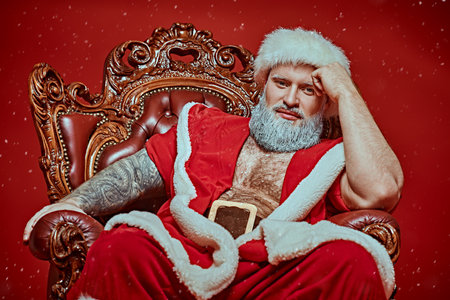 Christmas and New Year for adults. An attractive, muscular Santa Claus in a red fur coat sits relaxed in a chair and smiles at the camera. Red studio background with snowfall.の写真素材