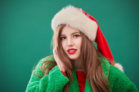 Merry Christmas and New Year! Happy pretty girl posing in a fashionable green fur coat and Santa Claus hat. Green studio background with copy space.の写真素材