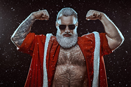 Strength and power. A handsome brutal muscular Santa Claus in a red fur coat and sunglasses stands covered in snow and looks at the camera. Black background with snowfall. Christmas and New Year.の写真素材