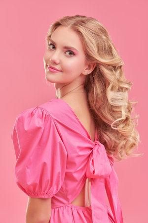 Feminine beauty. Portrait of a cute blonde girl with delicate pink makeup posing in an elegant pink dress on a pink studio background. Hairstyles, Hollywood wave.の写真素材