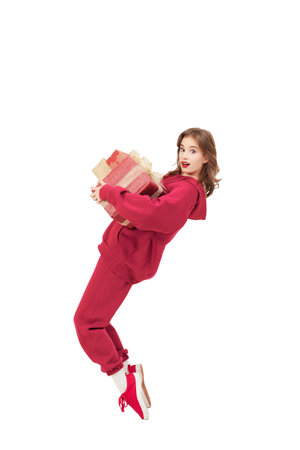 Christmas and New Year. Full-length portrait of a cheerful teenage girl,  wearing red sports suit, posing emotionally with a gift. White studio background.の写真素材